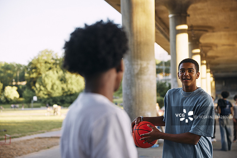 阳光明媚的一天，一个十几岁的男孩微笑着，手里拿着篮球，在桥下和朋友一起玩耍。图片素材