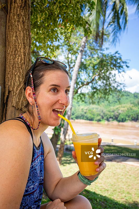 一位女士在湄公河畔的琅勃拉邦享受着清爽的热带芒果冰沙，度过夏日假期，她开心地笑着，感受着旅行和健康的生活方式图片素材