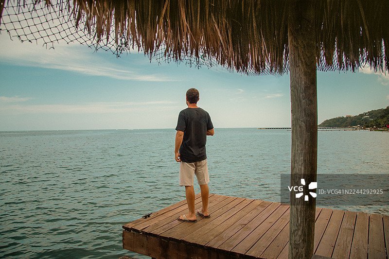 一个穿着休闲服的男人站在码头上，背对着镜头，望着洪都拉斯罗阿坦的加勒比海图片素材
