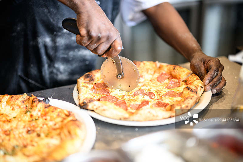 特写：在餐厅厨房的盘子里，用滚轮刀切开新鲜出炉、色彩鲜艳的意大利辣香肠披萨的手部特写图片素材