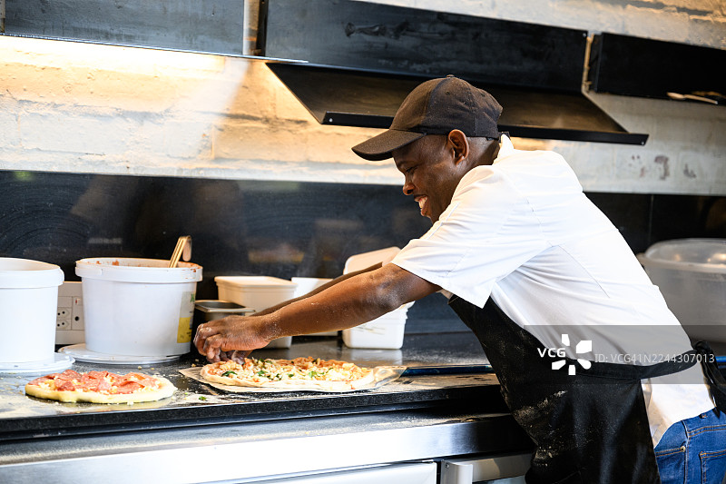 专业厨房里，一位快乐的厨师正在撒满面粉的台面上摆放未烹饪的比萨饼，侧面视角。图片素材