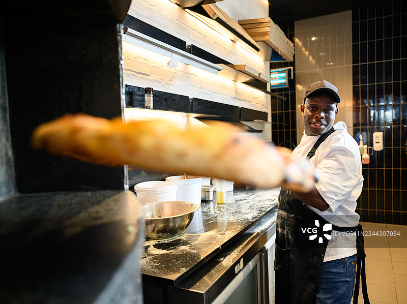 自信的黑人厨师在餐厅厨房里，从燃木烤箱中取出刚出炉的披萨图片素材