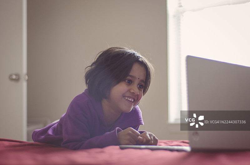 一个快乐的年轻印度女孩躺在红色的床上，脸上带着灿烂的笑容，正在看着一台笔记本电脑。图片素材