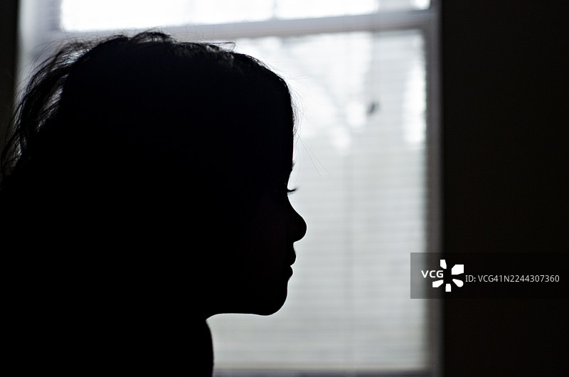 一位年轻女孩的黑白侧面剪影，目光投向明亮模糊的窗户。脸上的深邃阴影营造出沉思、希望或深刻思考的氛围。图片素材