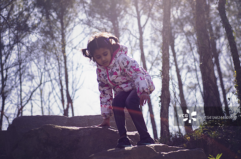 一个决心坚定的年轻印度女孩，穿着一件色彩鲜艳的图案冬季夹克，正在户外小心翼翼地在岩石上行走。图片素材