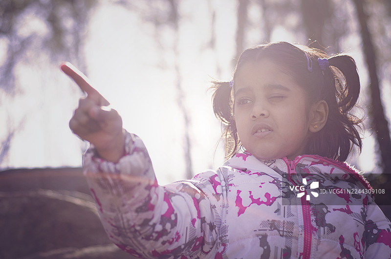 一个可爱的年轻女孩，穿着印花冬季夹克，脸上带着坚定或兴奋的表情，手指指向镜头外，明亮的逆光照在她脸上，让她微微眯起了眼睛。图片素材