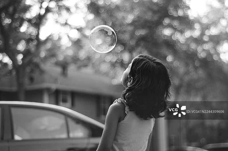 一张黑白照片，捕捉了一个安静的瞬间：一个小女孩穿着灰色上衣，注视着一个正在上升的、彩虹般闪耀的泡泡。图片素材