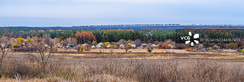 深秋时节的乡村景象，传统房屋点缀在色彩斑斓的落叶和常绿树木之间，前景是开阔的田野，远方是茂密的森林。图片素材