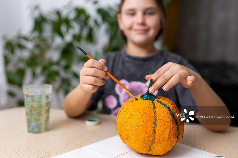 一个孩子正在用画笔给万圣节制作的亮橙色纸浆南瓜涂上颜色，南瓜还带着绿色的茎。图片素材