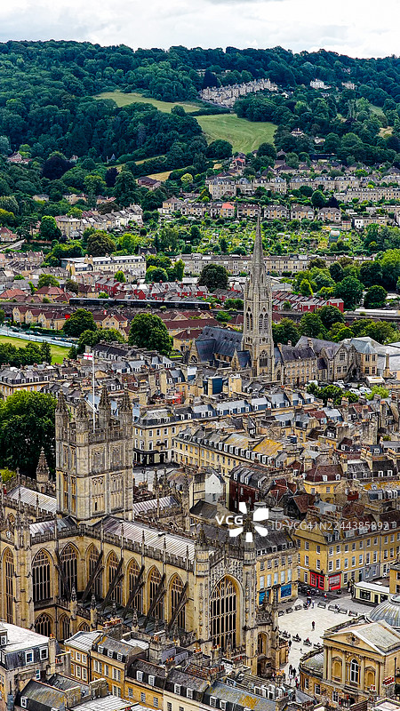 英格兰萨默塞特郡巴斯市巴斯修道院的鸟瞰图,有英国国旗、圣约翰福音传教士教堂尖塔和一列行驶的火车,周围环绕着乔治亚风格的建筑和乡村山丘,这是联合国教科文组织世界遗产地。图片素材