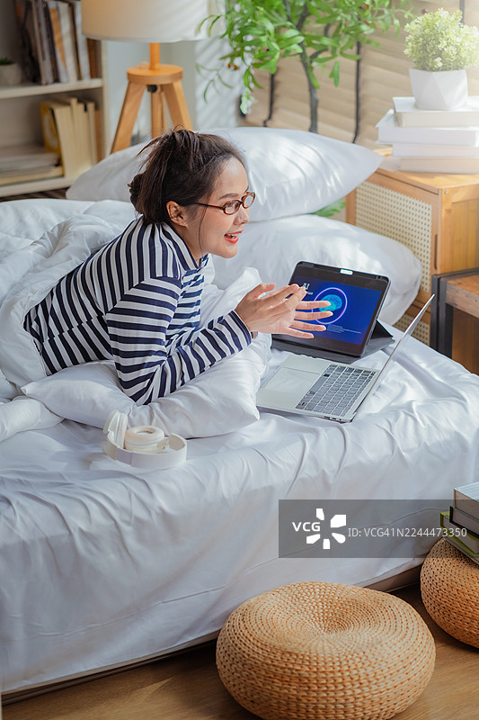 专业女性躺在床上，利用智能手机和平板电脑上的AI助手进行自动化工作流程优化，运用尖端机器学习技术提高业务生产力，在家进行自由职业分析和工作。图片素材