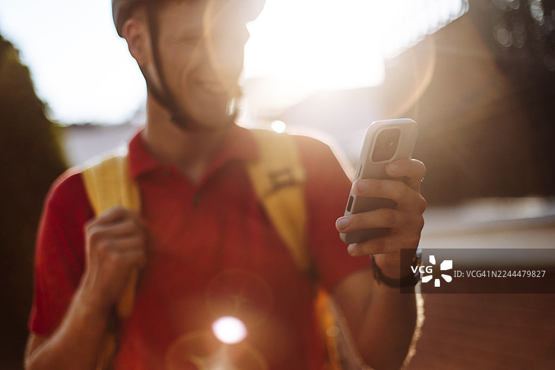 阳光明媚的户外，一个快乐的男人背着黄色背包，在明亮的阳光和镜头光晕下使用智能手机。图片素材