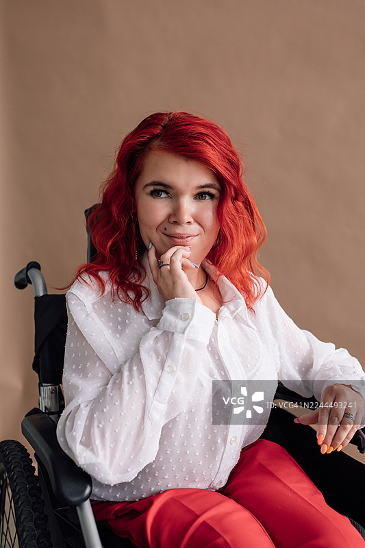 一位自信的红发女子坐在轮椅上，身穿白色波点衬衫和红色裤子，姿态沉思图片素材