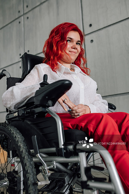 一位自信的女士坐在轮椅上，身穿白色衬衫和红色裤子，在现代环境中微笑。图片素材