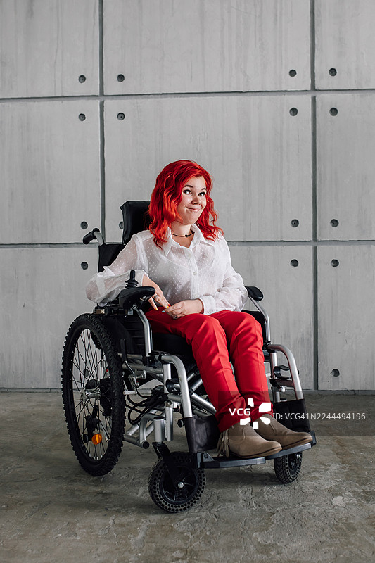 自信的女士坐在轮椅上，身穿白色衬衫和红色裤子，在现代水泥背景下微笑图片素材