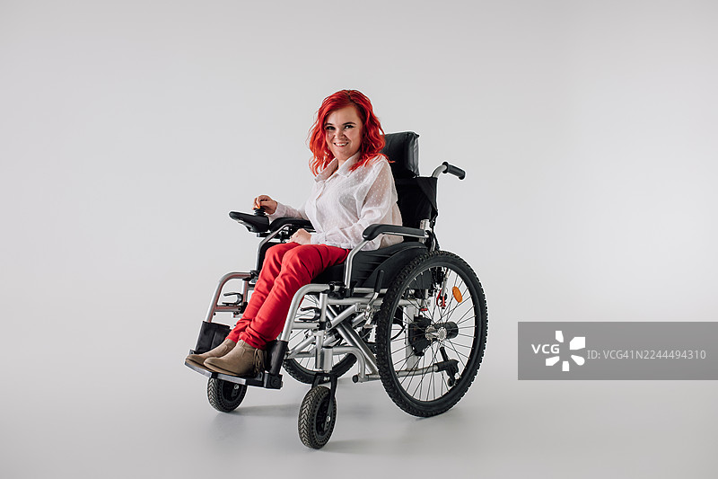 一位红发女子坐在轮椅上，穿着休闲的白色衬衫和红色裤子，在影棚里微笑图片素材