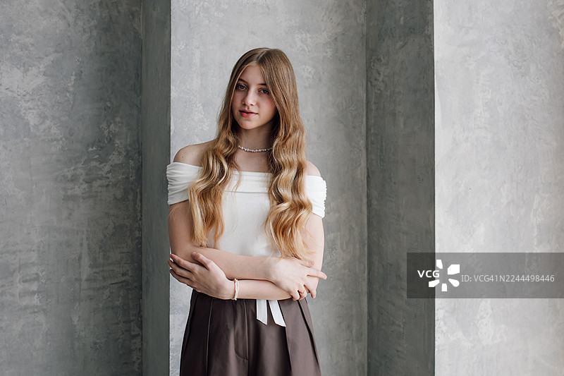 一位身穿露肩白色上衣的年轻女子，留着金色长发，背景是灰色混凝土墙图片素材