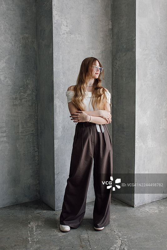 一位时尚女子身穿露肩白上衣和阔腿棕色裤子，站在混凝土背景前。图片素材