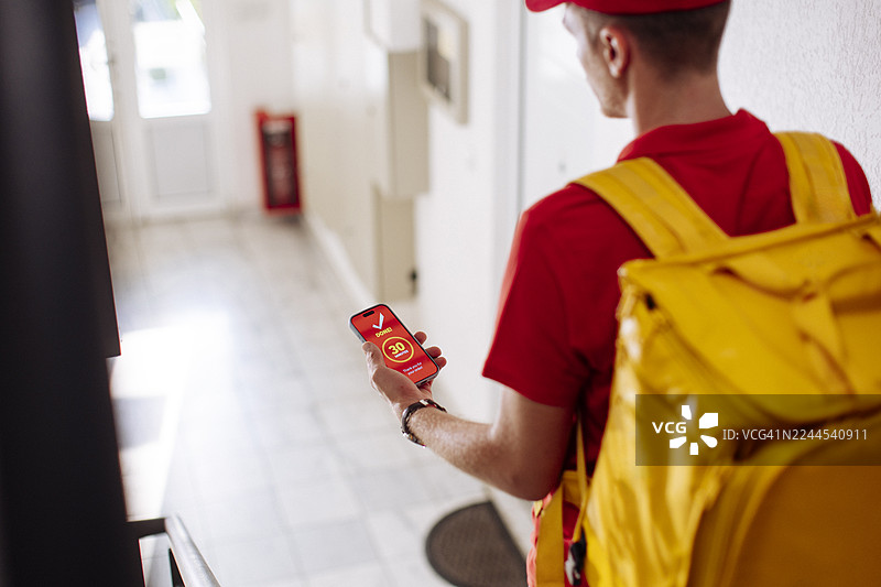 身穿红色制服、背着黄色背包的快递员在楼道里查看智能手机图片素材