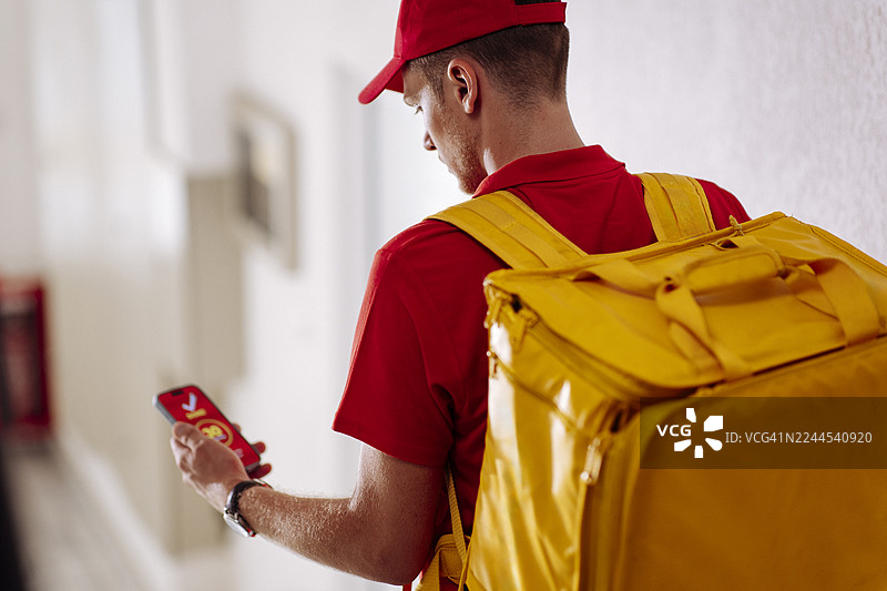 送货员背着黄色背包，在走廊里边走边看手机图片素材