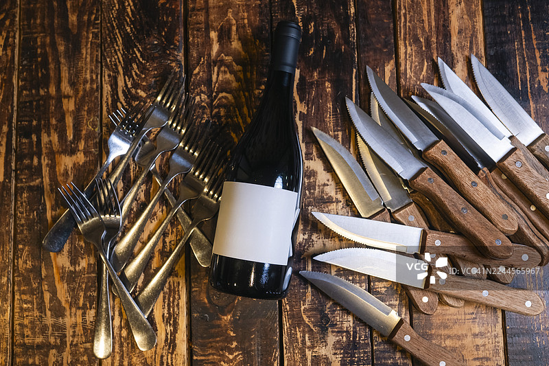 木质桌子上，酒瓶被叉子和刀子环绕。图片素材