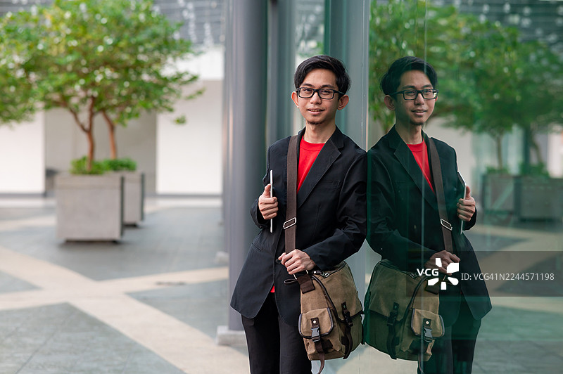 一位敬业的穆斯林商务高管，身背斜挎包，手持数字平板电脑，倚靠在反光玻璃结构旁拍摄的肖像照。图片素材