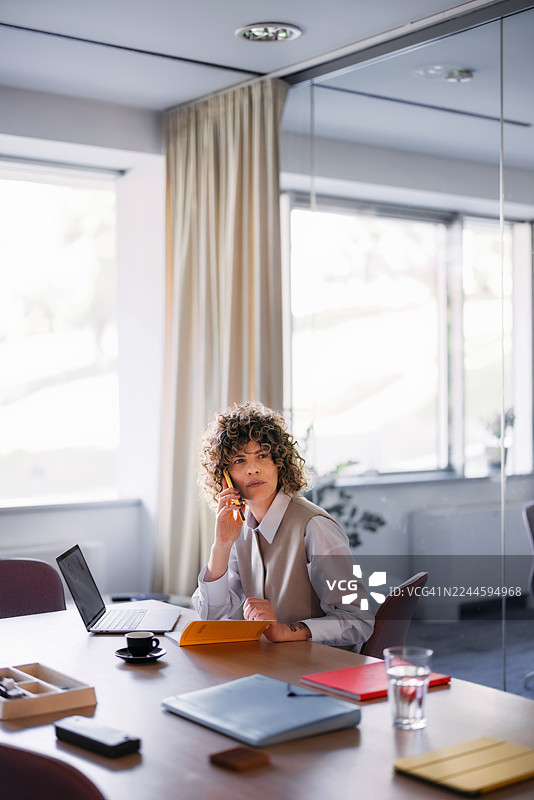 职业女性在办公室办公桌旁，一边使用笔记本电脑、记事本，一边接听电话，旁边还有一杯水。图片素材