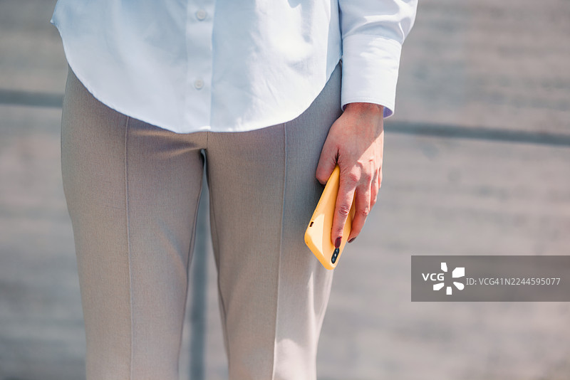 手持黄色智能手机的特写，身穿白衬衫和米色裤子，展现休闲专业的造型图片素材