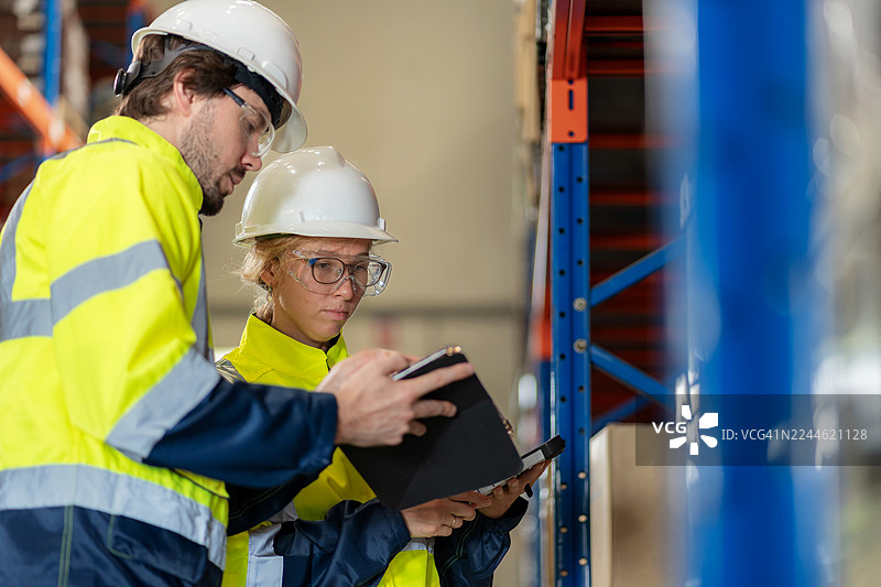 两位穿着反光安全服的分销仓库员工在库存检查期间合作。一名女工正在使用条形码阅读器扫描箱子，而一名男同事则在数字平板电脑上查看数据。图片素材