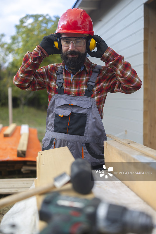 留着胡子的男性木匠，身穿安全装备，在建筑工地上调整耳罩。图片素材