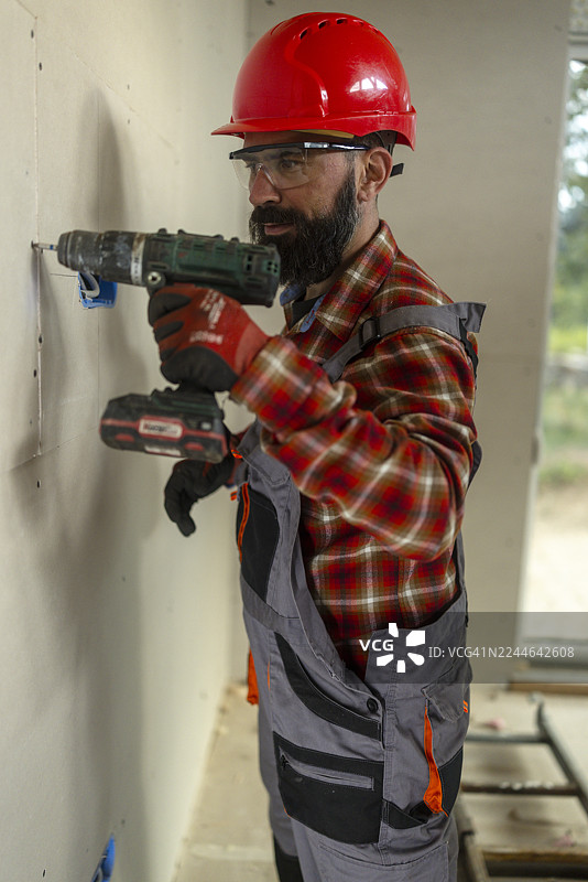 正在建造的木屋中，一位留着胡子的男性建筑工人戴着红色的安全帽正在钻墙。图片素材