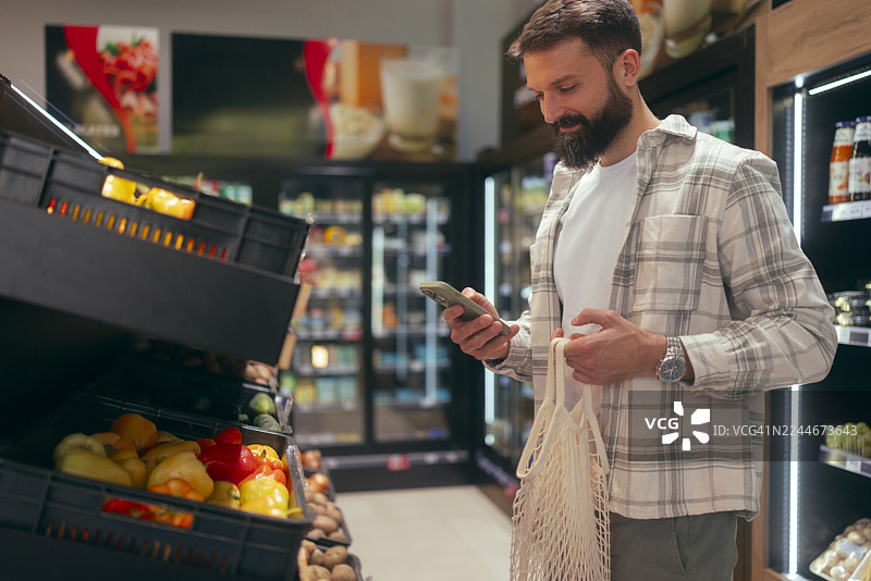 现代男子在超市用手机查看价格或营养信息，同时从货箱中挑选蔬菜。图片素材
