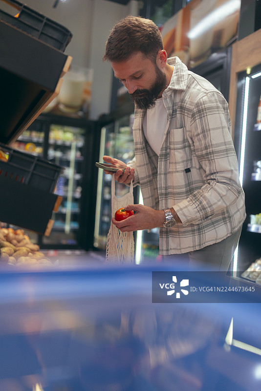 一位休闲男子站在生鲜区，一手拿着蔬菜和手机，另一手提着环保购物袋。图片素材