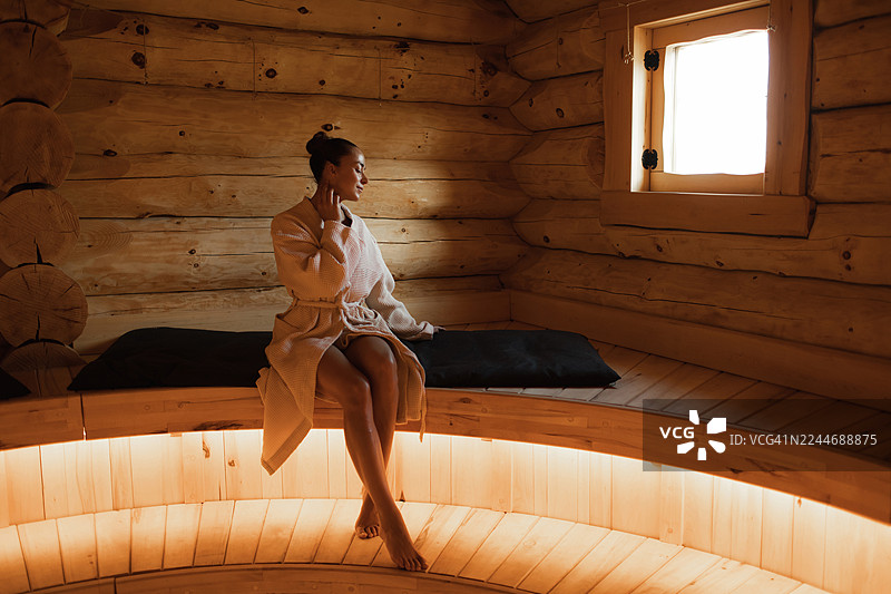 舒适的女士穿着浴袍坐在温暖的木制桑拿房窗边图片素材