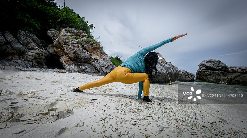 一位女士在热带海滩上练习瑜伽，背景是岩石悬崖，画面传达出放松、平衡与亲近自然的意境。图片素材
