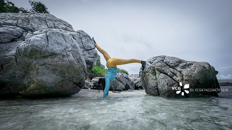 女子在热带海滩上练习瑜伽，背景有岩石悬崖，画面传达出放松、平衡与亲近自然的意境。图片素材