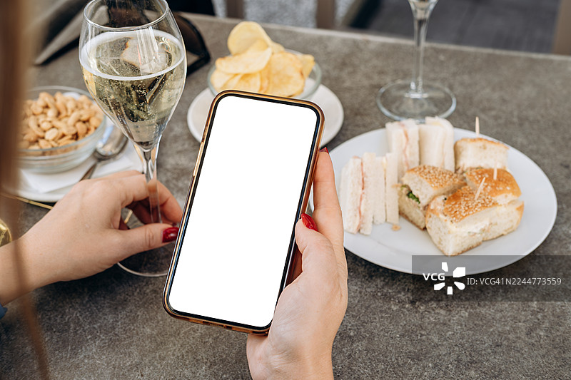 在咖啡馆场景中，女子手持带有空白屏幕的智能手机模型，周围环绕着精致的饮品和美味的开胃菜，非常适合设计使用图片素材