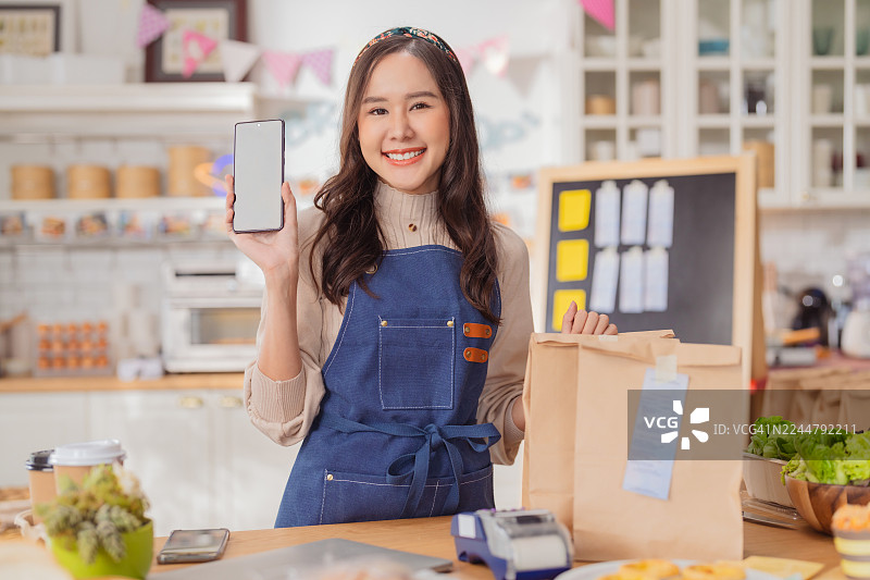 一位快乐的小咖啡馆店主微笑着展示智能手机屏幕，准备通过数字订购系统接受在线订单和配送服务，以推广其小型烘焙面包业务的肖像照图片素材