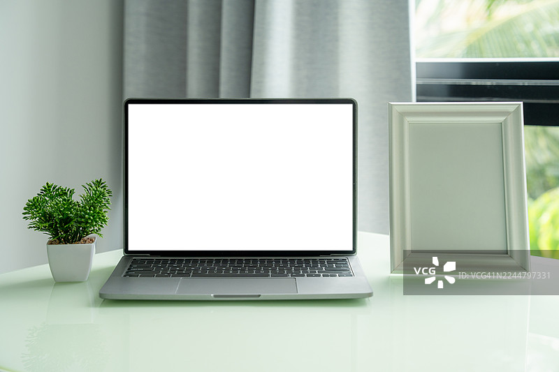 在家办公的场所，桌上放着笔记本电脑。笔记本电脑的屏幕是白色的空白屏，放在木制工作台上，旁边还有相框和盆栽，在一个有装饰的极简办公室里。图片素材