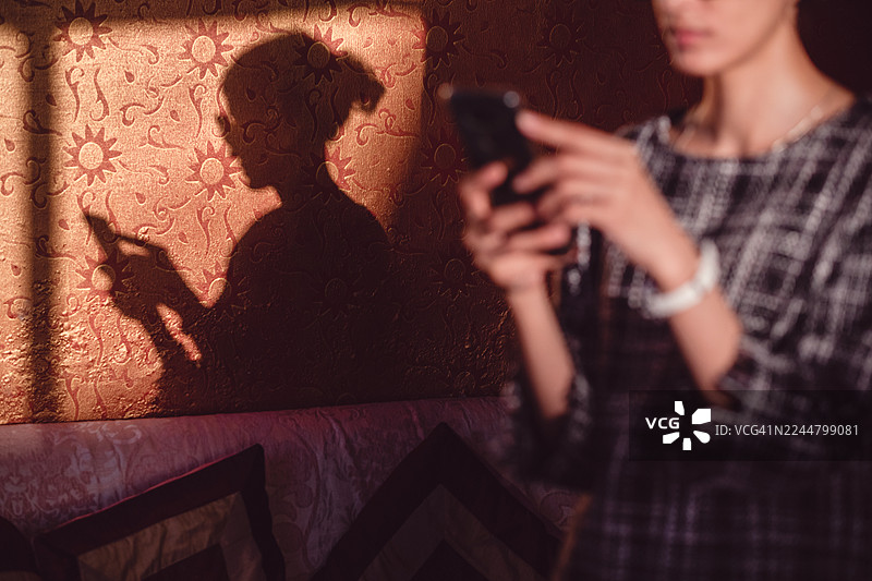 强烈的午后阳光照射下，一位女士使用手机的深色侧影投射在室内有图案的橙色墙壁上。图片素材