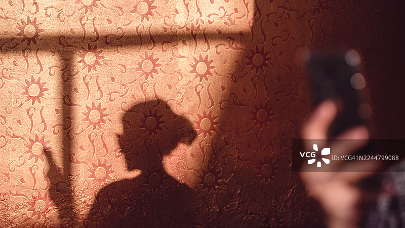 强烈的午后阳光照射下，一名女子使用手机的深色侧影投射在室内带有图案的橙色墙壁上。图片素材