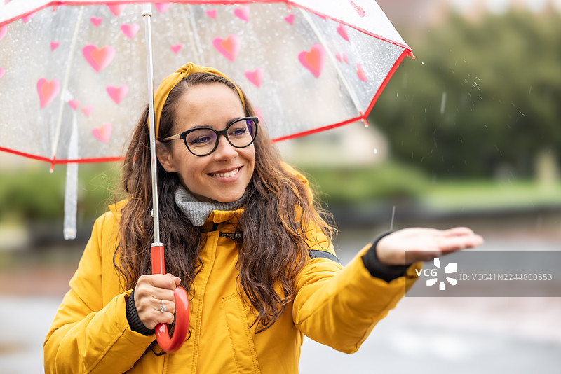一位年轻女子穿着黄色带帽防水夹克，撑着伞，面带微笑，张开双臂，尽情享受雨天和新鲜空气。图片素材