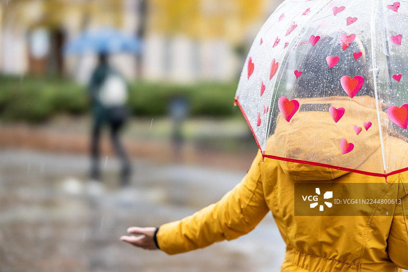 一名身穿黄色连帽防水夹克的女子背影，她撑着伞，伸出手，仿佛在确认是否还在下雨。图片素材