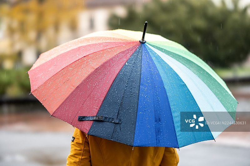 穿着黄色夹克的人撑着一把彩虹色的大雨伞，雨滴落下，明显显示出深秋的雨天景象。图片素材