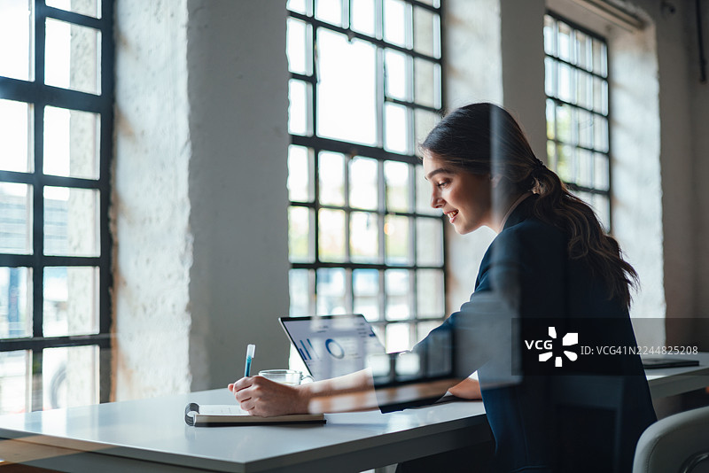 职业女性在现代办公室的办公桌上使用笔记本电脑，旁边放着笔记本和带有大格子的窗户图片素材