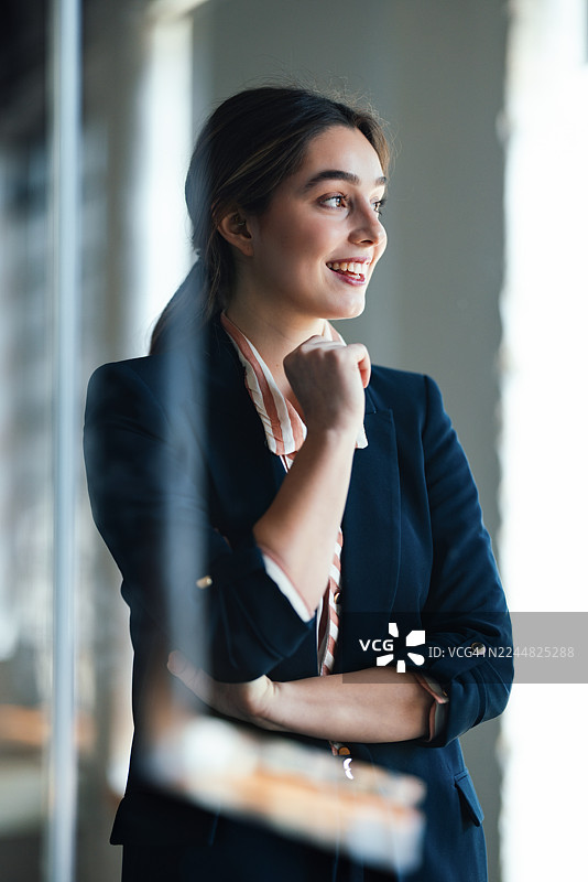 自信的女商人在现代西装外套中，在窗边微笑，展现出专业、充满希望的工作场所瞬间图片素材