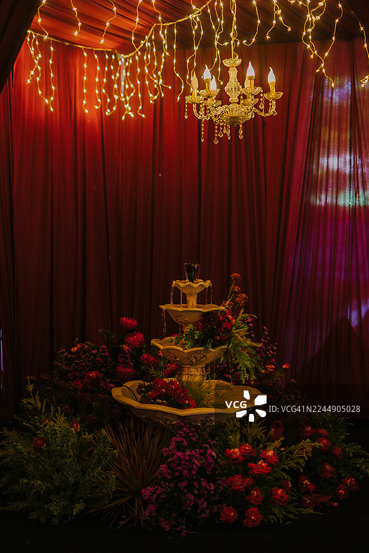 一场高端活动中的装饰性多层喷泉和鲜花，配有红色帷幕、吊灯和温暖的串灯。图片素材