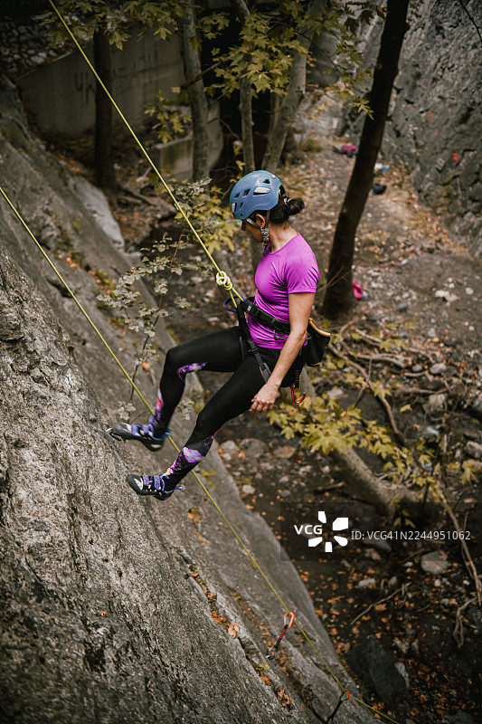 身穿紫色T恤、戴着头盔和安全带的女性攀岩者在户外悬崖上进行下降。图片素材