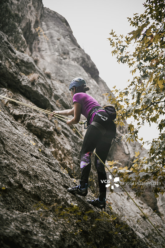 身穿紫色T恤的女性攀岩者在雨中，借助绳索装备攀爬岩石峭壁图片素材