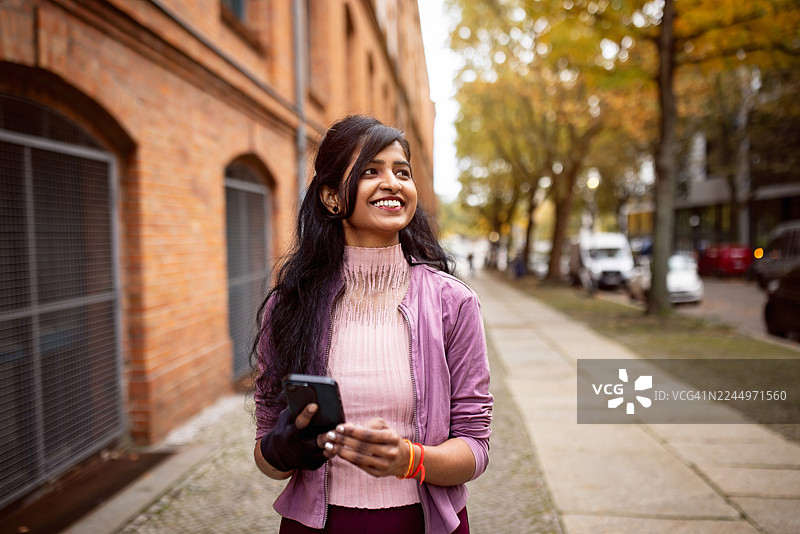 一位年轻的印度女性走在柏林街头，靠着砖墙，面带微笑地拿着智能手机图片素材